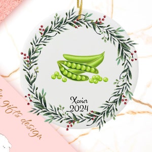 May include: White ceramic ornament with a green wreath and red berries. The center features a watercolor illustration of green peas in a pod with the text "Xavier 2024".