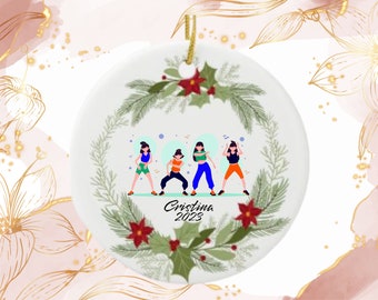 May include: White ceramic ornament with a gold string for hanging. The ornament features a design of four dancing figures in colorful outfits, surrounded by a wreath of green leaves, red berries, and the text "Cristina 2023".