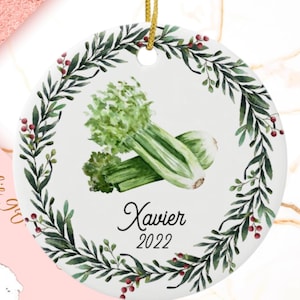 May include: A white ceramic Christmas ornament with a green leafy wreath and red berries. In the center is a watercolor illustration of celery with the name "Xavier" and the year "2022" written below.