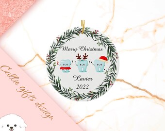 May include: A white round Christmas ornament with a green wreath border. The ornament features three cartoon teeth with red and white accents. The text "Merry Christmas" and "Xavier 2022" is printed on the ornament.
