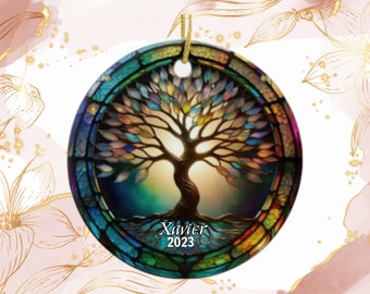 May include: A round, stained-glass style ornament featuring a tree design with colorful leaves and a brown trunk. The ornament has a gold string for hanging and the name "Xavier" and the year "2023" are printed on the bottom.