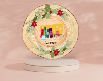 May include: A wooden book club ornament with a festive design. The ornament features a central graphic of books, a teacup, and the words "BOOK CLUB." The name "Xavier" and the year "2022" are also displayed. The ornament is decorated with holly and poinsettias.
