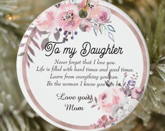 May include: A round, white ceramic ornament with a floral design in pink and purple hues. The text "To my Daughter" is displayed, along with a heartfelt message from "Mom". A gold string is attached for hanging.