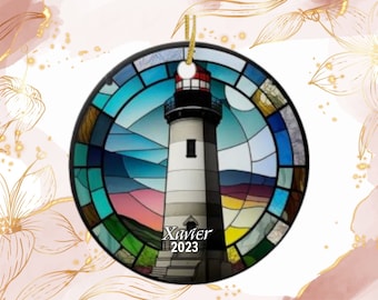 May include: Round ceramic ornament featuring a lighthouse design. The lighthouse is white with a red top, set against a stained-glass style background of blues, greens, and pinks. The name "Xavier" and the year "2023" are printed on the ornament.