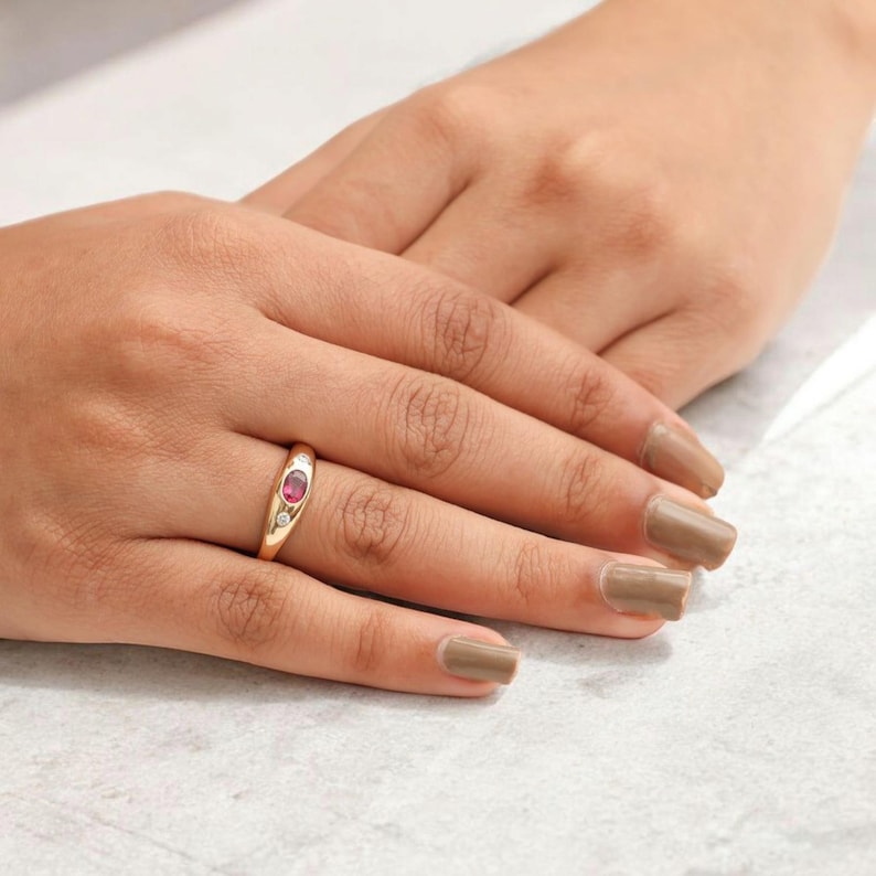 May include: A gold ring with a red oval gemstone and two smaller white gemstones. The ring is on a person's finger.