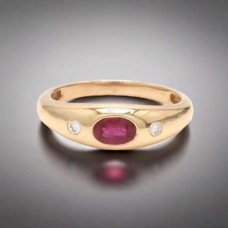 May include: A gold ring with a red ruby gemstone and two smaller diamonds. The ring has a smooth, rounded band.