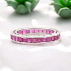 Könnte beinhalten: Ein silberner Ring mit einem durchgehenden Band aus quadratisch geschliffenen, leuchtend rosa Edelsteinen. Die Fassung des Rings ist aus hellem, poliertem Silber, und die Edelsteine liegen bündig mit der Oberfläche des Bandes. Ein Schmuckstück.