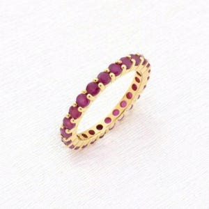Puede incluir: Un anillo de oro con una fila continua de piedras preciosas redondas de color rojo intenso. La banda del anillo es de color oro pulido, y las piedras preciosas están espaciadas uniformemente alrededor de toda la circunferencia. Sobre fondo blanco.