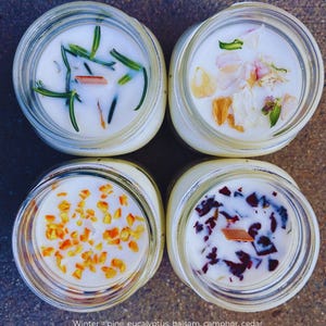 Op de afbeelding: Vier glazen potten met deksels, elk met een andere geurkaars. De kaarsen zijn versierd met verschillende planten en gedroogde bloemen. De potten zijn in een vierkant patroon gerangschikt. De tekst onder de potten luidt: "Winter = pine, eucalyptus, balsam, camphor, cedar Spring = lemongrass, lime, black currant, thyme, sage Summer = mandarin, lemon, grass, pineapple, vanilla Fall = ozone, amber, clove, pear, palo santo"