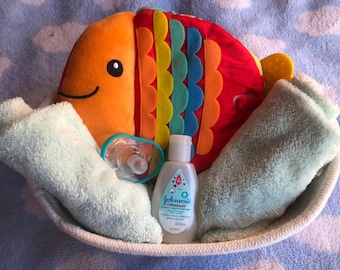 Puede incluir: Una canasta de regalo para bebés con un juguete de peluche de pez naranja, un chupete, un frasco de jabón para bebés Johnson's y dos toallitas de color azul claro. Los artículos están dispuestos en una cesta tejida blanca sobre una manta azul con estampado de nubes.