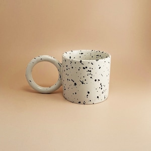 May include: A white ceramic mug with a black speckled design and a unique circular handle.