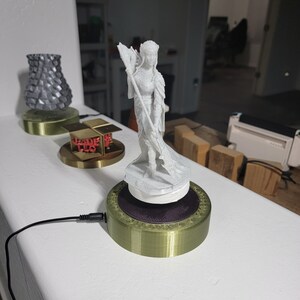 May include: A white 3D printed figurine of a woman holding a spear, standing on a rotating base with a gold and green finish. The base is connected to a black power cord.