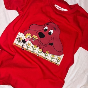 May include: A bright red t-shirt featuring a cartoon graphic of Clifford the Big Red Dog with a girl and flowers. The graphic shows Clifford with a girl and flowers. The shirt has a crew neck and short sleeves.