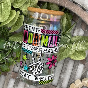 May include: A clear glass jar with a wooden lid, featuring the text "BEING NORMAL IS OVERRATED STAY WEIRD". The jar is decorated with a skeleton hand making a peace sign, stars, and a pink flower. The jar is sitting on a wooden tray.