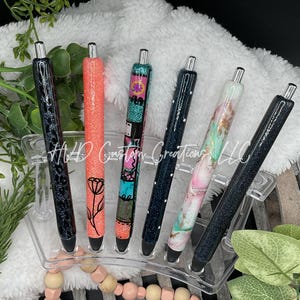 May include: A collection of six glitter pens in various colors and designs. The pens are displayed in a clear acrylic holder. The pens feature black, coral, teal, and floral patterns. The pens are a stylish writing accessory.
