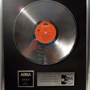 Könnte beinhalten: Eine silberne Schallplatte, die ABBA für ihr Album Greatest Hits verliehen wurde. Die Auszeichnung ist in einem schwarzen und silbernen Rahmen gerahmt und trägt den Text "Diese vergoldete Schallplatte wird in Anerkennung der prestigeträchtigen Leistung verliehen, den Platin-Status erreicht zu haben." Die Auszeichnung trägt auch das Logo der World Wide Music Awards.