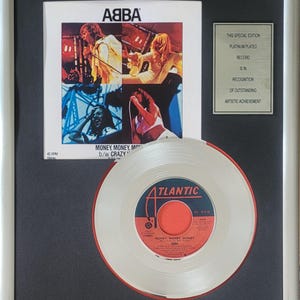 Könnte beinhalten: Gerahmte, platinbeschichtete Schallplatte der ABBA-Single "Money, Money, Money". Die Schallplatte ist weiß mit einem roten Mittellabel und dem Atlantic-Logo. Der Rahmen enthält das Albumcover und eine Plakette mit der Aufschrift "This Special Edition Record Is In Recognition Of Outstanding Artistic Achievement."