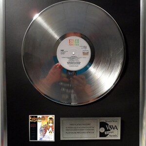 Op de afbeelding: Een zilveren platenplaat in een zwart frame met een zilveren plaat erin. De plaat is een platina-award voor de soundtrack van Labyrinth van David Bowie. De award wordt uitgereikt door EMI America en World Wide Awards Europe.