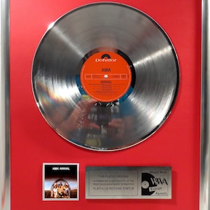 Könnte beinhalten: Eine silberne Platin-Schallplatte für das Album Arrival von ABBA. Die Schallplatte ist gerahmt und auf einem roten Hintergrund ausgestellt. Die Auszeichnung wird von den World Wide Music Awards Europe verliehen.