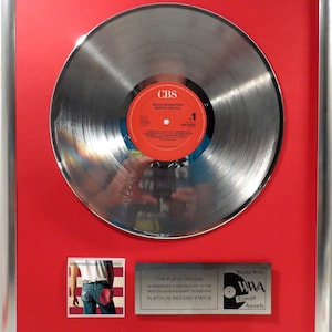 Puede incluir: Un disco de platino plateado para el álbum de Bruce Springsteen "Born in the USA" con un fondo rojo y una etiqueta CBS. El premio es otorgado por los World Wide Music Awards Europe.