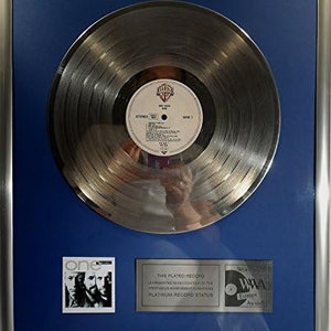 Puede incluir: Un disco de platino plateado con fondo azul. El disco está enmarcado y tiene una etiqueta con el texto "One" y una foto de grupo de tres hombres. El texto "This plated record is presented in recognition of the prestigious achievement in recording platinum record status" está en una placa debajo del disco. El texto "World Music Awards" está en una placa debajo del disco.