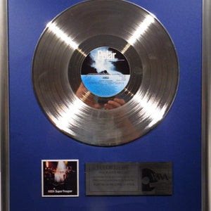 Könnte beinhalten: Eine silberne Platin-Schallplatte für das Album "Super Trouper" von ABBA. Die Schallplatte ist in einem blauen Passepartout und einem silbernen Rahmen gerahmt. Die Schallplatte ist von einem silbernen Rand umgeben und trägt ein Etikett mit dem Namen der Band und dem Titel des Albums. Die Auszeichnung enthält auch ein kleines Foto des Albumcovers und eine Plakette mit der Aufschrift "Diese plattierte Schallplatte bezeugt eine Multi-Platin-Aufnahmeleistung. Platin-Status."