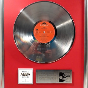 Op de afbeelding: Een zilveren platenplaquette met een rode achtergrond. De plaquette is ingelijst en heeft een zilveren plaat met een label waarop "Polydor ABBA More ABBA Gold" staat. De plaquette bevat ook de tekst "Deze geplateerde plaat wordt uitgereikt ter erkenning van de prestigieuze prestatie om de platina-status te bereiken" en "World Wide WWA Europe Awards".