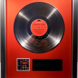 Könnte beinhalten: Ein silbernes Schallplattenalbum, gerahmt in einem silbernen Rahmen mit einem roten Passepartout. Das Album trägt den Titel "The Very Best of ABBA" und ein Polydor-Label. Das Album ist mit einem kleinen Bild des Albumcovers und einer Plakette gerahmt, auf der steht "This Platinum Record is presented in recognition of over 1,000,000 sales in the UK."