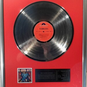 Op de afbeelding: Ingelijste platina plaat van ABBA's "16 ABBA-HITS" album. De plaat is zwart met een rood en wit label. De lijst is zilverkleurig met een rode achtergrond. Inclusief een kleine albumhoesafbeelding en een plaquette.