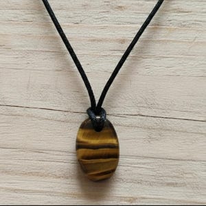Op de afbeelding: Een ketting met een tijgeroog hanger. De ovale hanger is een gepolijste, goudbruine steen met horizontale banden. Hij hangt aan een zwart koord, bovenaan geknoopt. De achtergrond is een licht houten oppervlak.