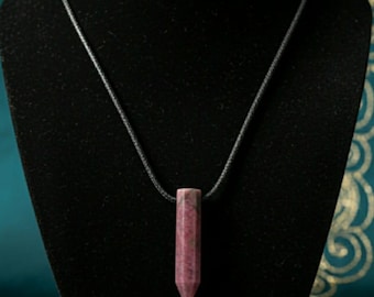 May include: A pink and maroon crystal pendant necklace. The pendant is a rectangular shape with a pointed bottom. The necklace is displayed on a black velvet bust against a teal background.