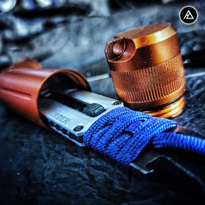 May include: A close-up of a silver Gerber multi-tool with a blue paracord wrap. The tool is partially open, revealing a blade and a small, silver button. A copper-colored bottle cap is visible in the background.