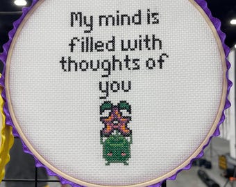May include: A white embroidery hoop with purple ribbon trim, featuring the text "My mind is filled with thoughts of you" and a pixelated image of a green and purple plant.