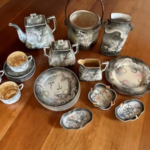 May include: A collection of antique Japanese porcelain tea set pieces. The set includes a teapot, sugar bowl, creamer, cups, saucers, plates, and serving dishes. Each piece is decorated with a dragon motif in shades of gray and white.