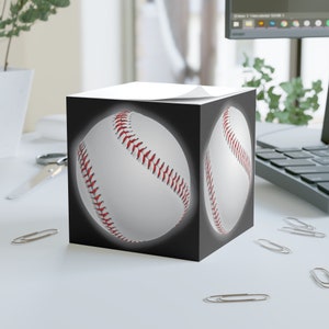 Könnte beinhalten: Ein schwarzer und weißer würfelförmiger Notizblock mit einem Baseball-Motiv auf jeder Seite. Der Baseball ist weiß mit roten Nähten.