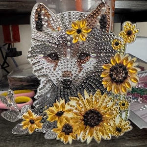 May include: A decorative piece featuring a wolf's head adorned with sparkling rhinestones. The wolf is surrounded by yellow and brown sunflowers. The piece has a clear, reflective surface, and the sunflowers add a touch of nature-inspired charm.