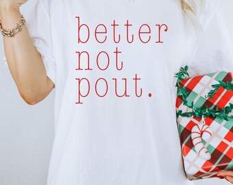 May include: White t-shirt with the red text "better not pout." A wrapped gift with red, green, and white plaid paper and candy cane accents is held in the hand. The shirt is a casual style.