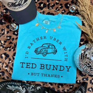 May include: A turquoise Comfort Colors t-shirt with the text "I'd rather Uber with Ted Bundy, but thanks." A black line drawing of a vintage car is centered on the shirt. A black and white baseball cap and gold necklace are also in the image.