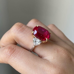 Op de afbeelding: Een gouden ring met een grote ovale robijn en twee kleinere driehoekige diamanten aan weerszijden.