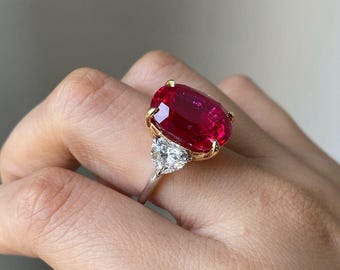 May include: A gold ring featuring a large oval ruby gemstone, flanked by two smaller triangular diamonds.