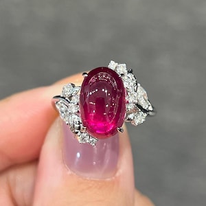Puede incluir: Un anillo de plata con un gran rubí cabujón de forma ovalada engastado en un delicado diseño de hojas. El rubí es de color rojo intenso y está rodeado de diamantes más pequeños.