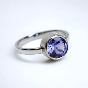 Puede incluir: Un anillo de plata con una gema grande, redonda y de color púrpura claro. La gema está engastada en un bisel y la banda es lisa y pulida. El anillo se muestra sobre un fondo blanco.