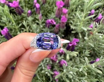 May include: A large, rectangular, blue gemstone ring with a halo of small, clear stones. The ring is set against a backdrop of green foliage and purple flowers.