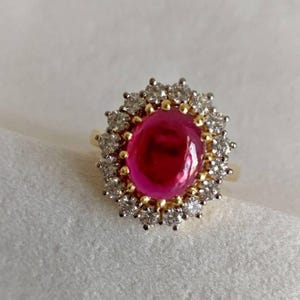 May include: A gold ring featuring a large, cabochon-cut ruby set within a halo of smaller diamonds.