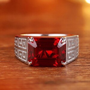 Puede incluir: Un anillo plateado con una gran gema rectangular de color rojo intenso. La banda del anillo presenta un patrón geométrico repetitivo. La gema es el punto focal, reflejando la luz.