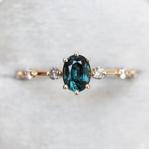 Può includere: Un anello d'oro con una pietra ovale verde scuro circondata da diamanti più piccoli. L'anello ha una fascia delicata con un design floreale.