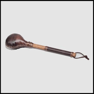 May include: A handcrafted percussion instrument, likely a rattle or shaker, featuring a rounded, leather-covered head and a wooden handle. The handle is wrapped with dark brown leather, and a leather loop is attached at the end. The instrument is designed for musical performance.