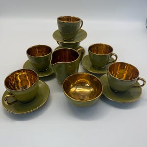 May include: A tea set comprising green teacups, saucers, a creamer jug, and a bowl. The interiors of the cups and bowl are gold. The items are arranged on a white surface.