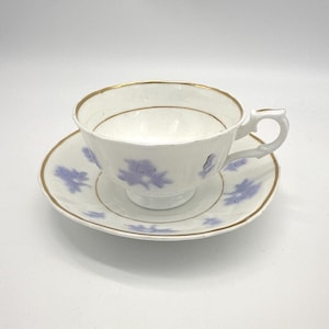 May include: White teacup and saucer set with a gold rim and delicate blue floral pattern. The teacup has a handle and sits on a matching saucer. A classic design for serving tea or coffee.