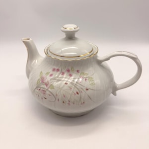 Puede incluir: Una tetera de cerámica blanca con tapa y asa. La tetera está decorada con diseños florales rosas y verdes y ribetes dorados. Un diseño clásico para servir té.